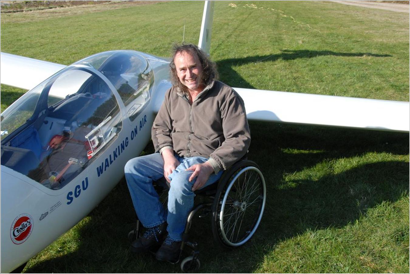 A middle aged gentleman with long curly hair sits in his wheelchair next to the WA1 glider. He has a very large smile on his face